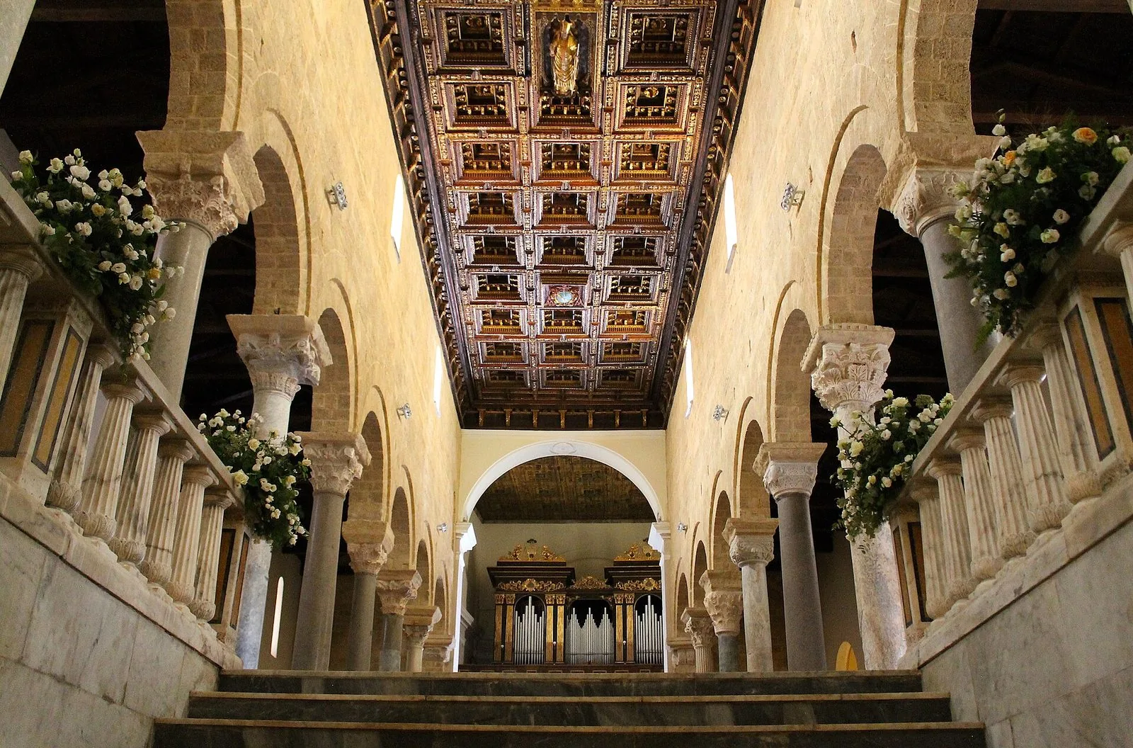 Navata centrale della Cattedrale di San Cataldo a Taranto, con colonne, archi, soffitto a cassettoni dorato e fiori bianchi lungo le balaustre
