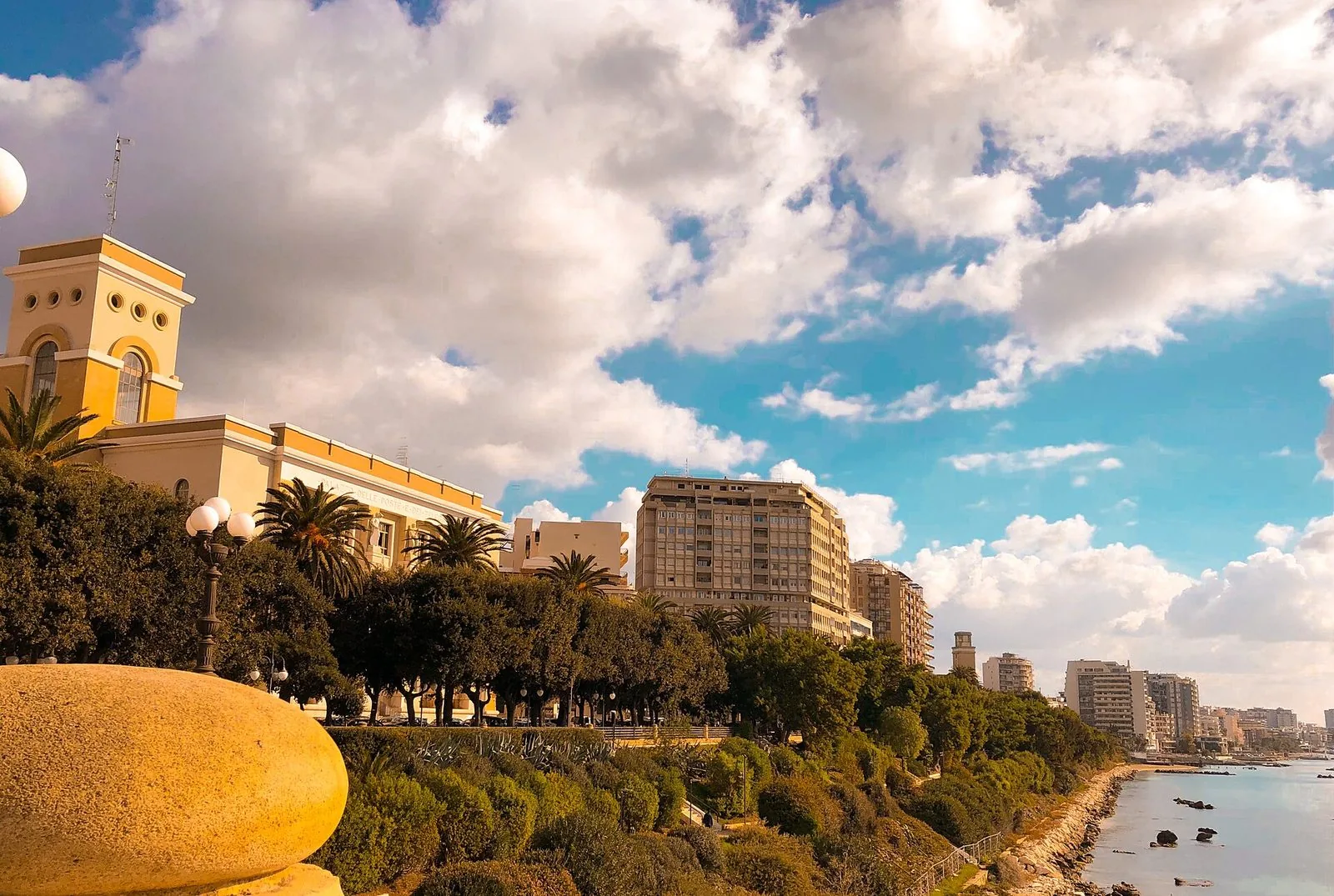 Tratto del Lungomare Vittorio Emanuele III a Taranto con il Palazzo delle Poste, palme e alberi, e il mare sulla destra sotto un cielo con nuvole