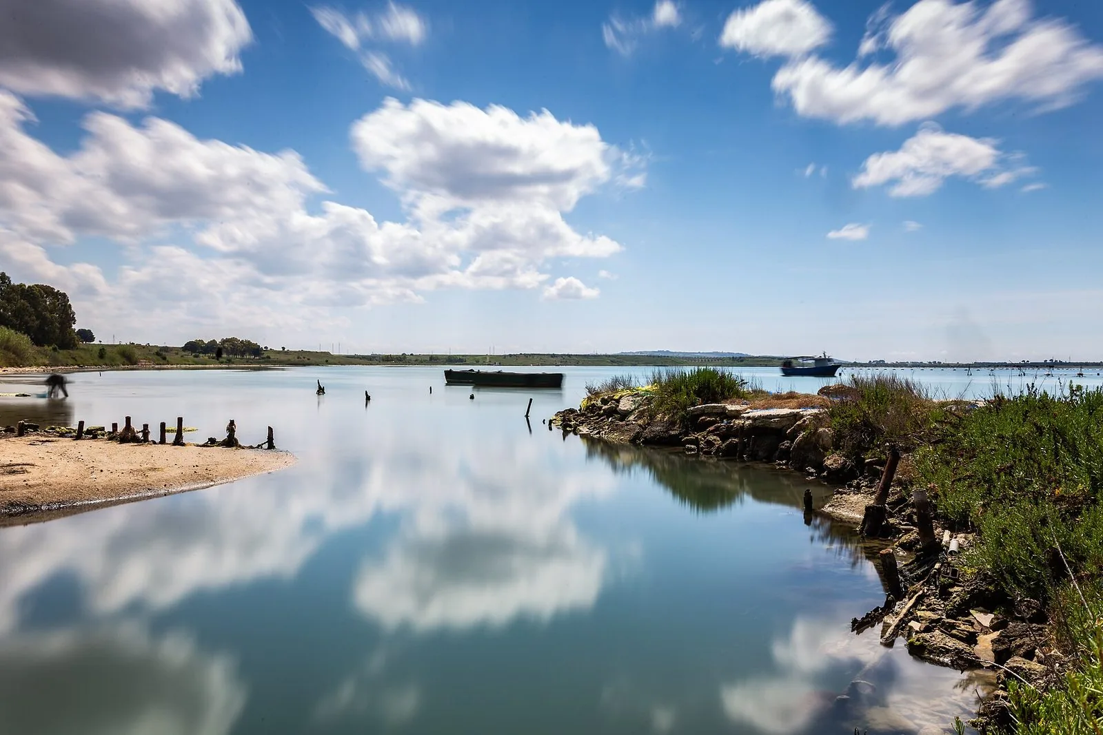 Sponda del Mar Piccolo di Taranto con barche ormeggiate, vecchi pali di legno nell'acqua e nuvole riflesse sulla superficie calma