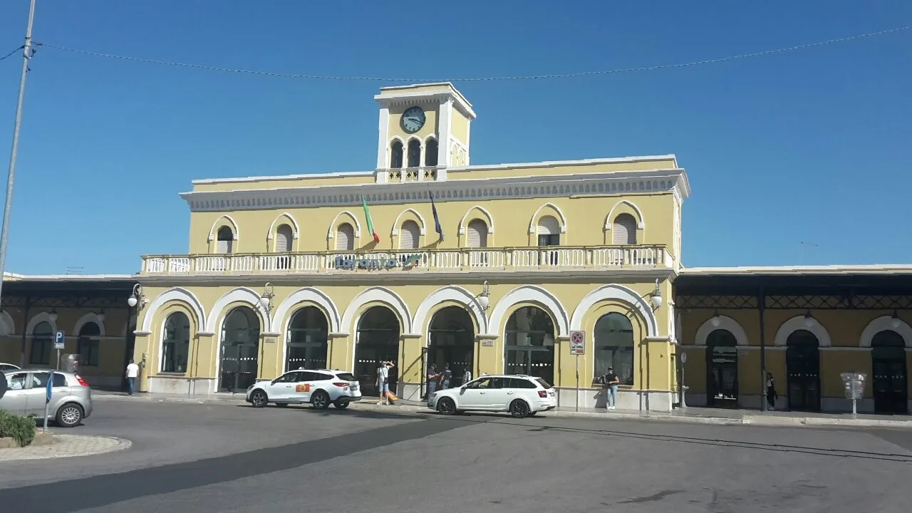 Facciata gialla della stazione ferroviaria di Taranto con torre dell'orologio, archi al piano terra e auto parcheggiate davanti